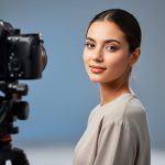Model in a modern studio portrait session with a Sony camera on a tripod softly blurred in the foreground and soft bokeh background lighting gear visible.